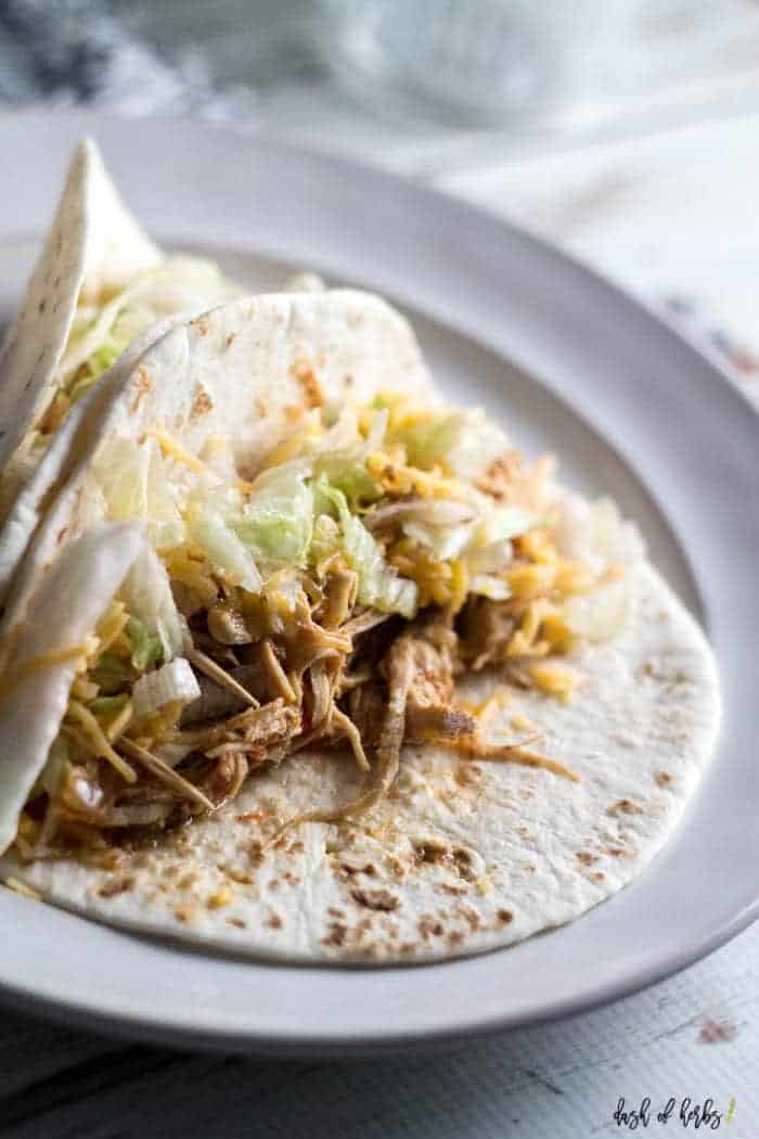 A close up image of the Instant Pot Easy Chicken Tacos recipe. There is one taco in the front of the picture, and 2 tacos together on a plate behind it. There is an empty bowl and navy blue napkin in the image as well.